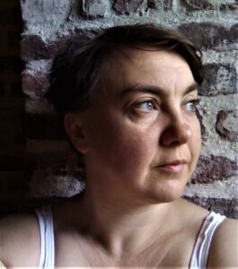 Stephe leans against a brick wall, her face turned towards the light from a nearby window.